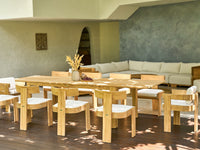 FORMA DINING TABLE set in a bright outdoor patio featuring natural teak wood with cushioned seats, surrounded by a cozy white sectional sofa and minimalist decor.