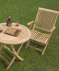 An outdoor setup with a teak bistro table and chair on lush green grass, elegantly set with a chessboard and drinks, inviting leisure and relaxation in a serene setting.