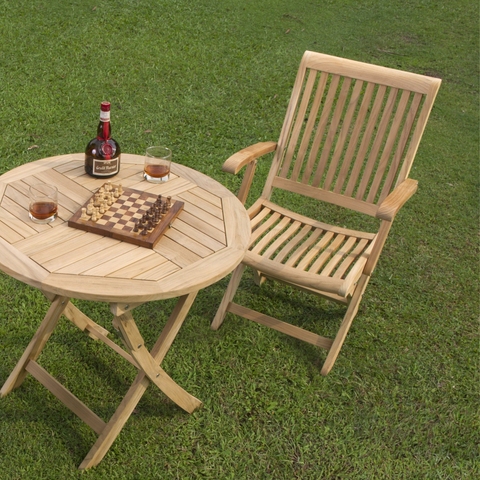 An outdoor setup with a teak bistro table and chair on lush green grass, elegantly set with a chessboard and drinks, inviting leisure and relaxation in a serene setting.