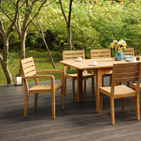 Outdoor dining setup with a teak table and matching chairs on a wooden patio, surrounded by lush greenery and decorated with a vase of flowers and cups.