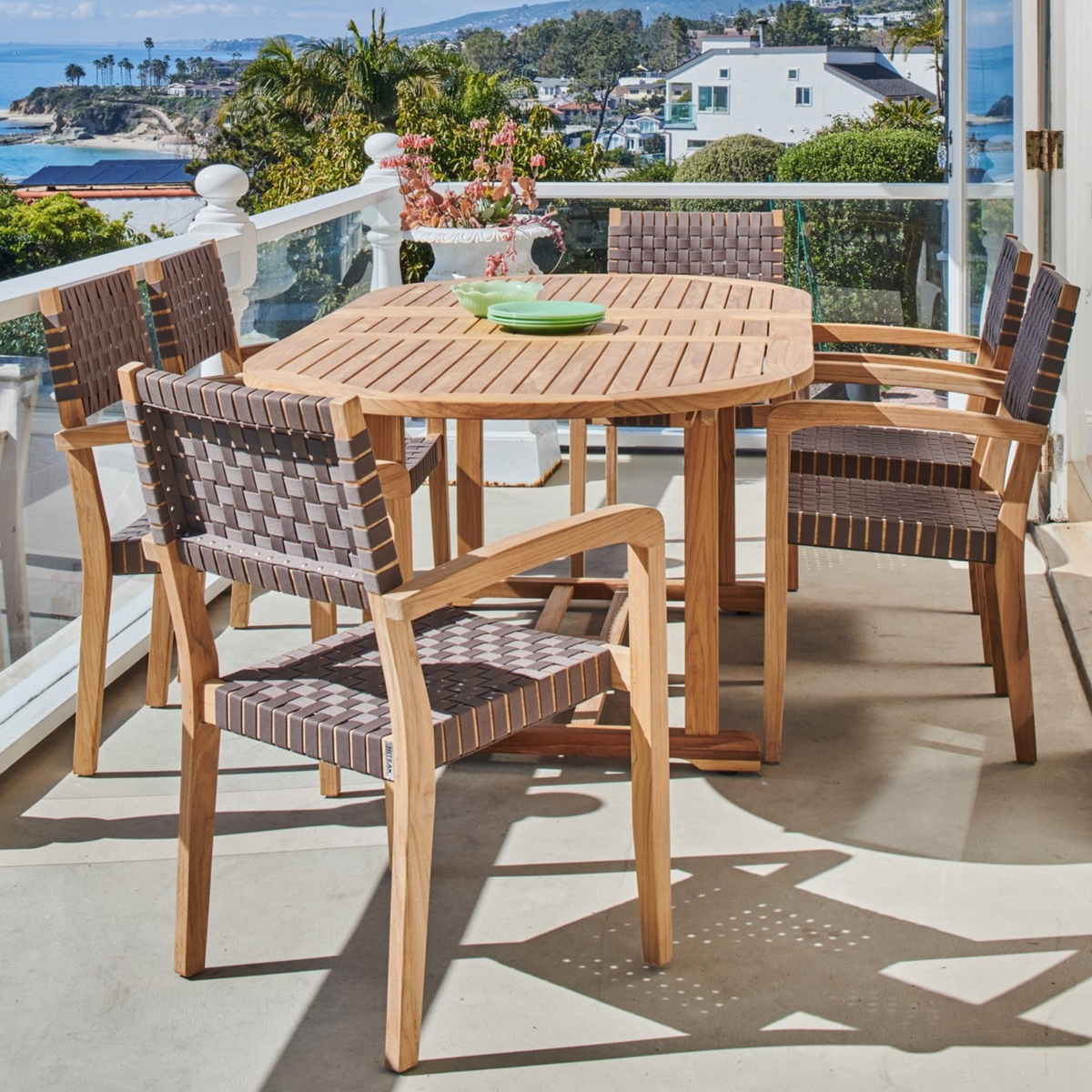 An inviting balcony setup with a teak dining table and woven chairs, overlooking a scenic coastal view for a serene dining experience.