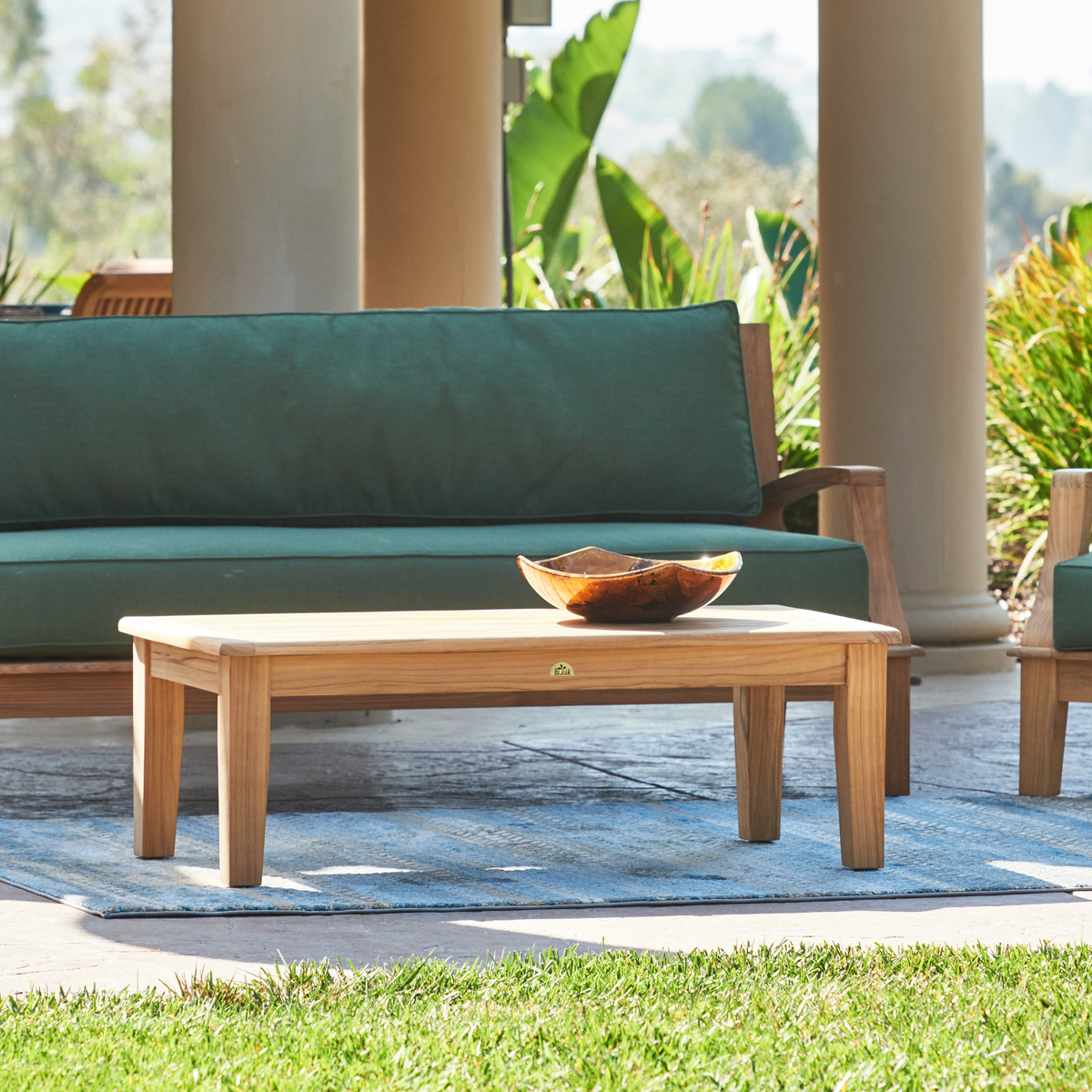 Outdoor lounge setting featuring a rectangular teak coffee table paired with green-cushioned teak furniture, placed on a patio surrounded by greenery.
